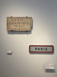 L'image est une photo prise dans le musée Carnavalet - Histoire de Paris avec une stèle gravée indiquant les limites de la ville et une plaque moderne de signalisation de l'entrée de Paris.