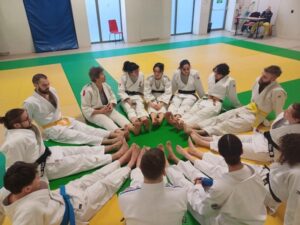 Les judokas sont assis, jambe tendu, de manière à faire un cercle. on me voit tout on fond, assise à une table.