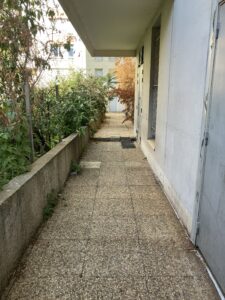 Une allée pavée de dalles en béton, 1,5 m de large. À droite, le mur d'un immeuble et la porte en métal d'un local poubelle, au dessus le plancher d'un balcon, à gauche la végétation d'un square déborde d'un muret. Au milieu du passage, deux parpaings. Au fond, une autre porte de local poubelle. On devine un espace un peu plus large.
